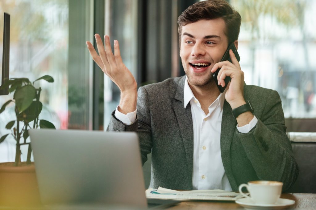happy businessman sitting by table cafe with laptop computer talking by smartphone while looking away (1)